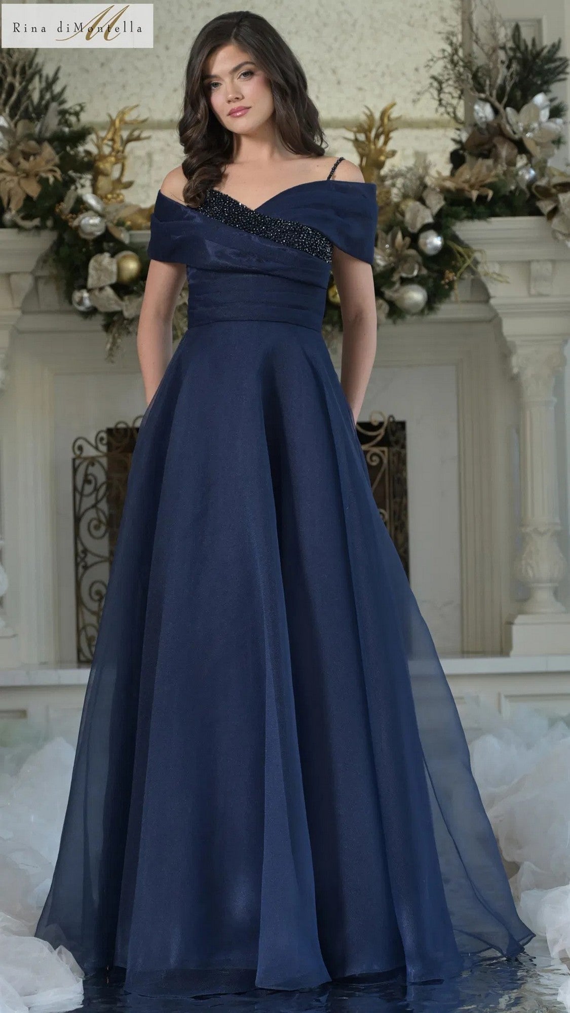 A woman poses indoors in front of a decorated fireplace wearing the navy Rina Di Montella RD3303, a long formal gown with an off-shoulder neckline and beaded details.